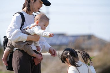 30 'lu yaşlarda Japon bir anne çocuklarıyla birlikte kırsalda oturuyor: 6 aylık bir erkek çocuğu ve yanında 3 yaşında bir kız çocuğu, huzurlu bir Mart öğleden sonra sarı çiçeklerin tadını çıkarıyor..
