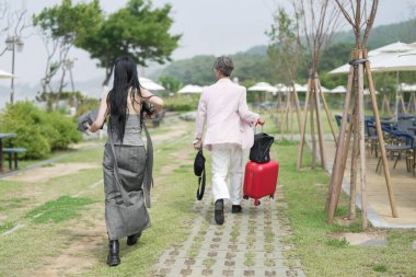In June 2025, a Korean man in his late 50s walks ahead with a suitcase, followed by a woman of the same age, as they pass a stylish seaside cafe terrace with tables, chairs, and umbrellas in Incheon.