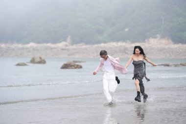 In June 2025, a Korean man and woman in their late fifties are running at full speed along the rugged shoreline filled with jagged rocks and crashing waves in Incheon Metropolitan City, South Korea.