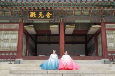 20 'li yaşlarda, pembe ve gökyüzü mavisi Hanbok giyen iki Koreli kadın, Haziran 2025' te Seul 'de Gyeongbokgung Sarayı' nın ön cephesinde oturuyorlar..