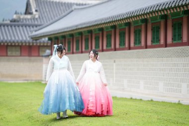 Yirmili yaşlarda iki Koreli kadın Gyeongbokgung Sarayı 'ndaki tarihi bir salonun önünde, açık bir yaz gökyüzü altında, pembe ve gökyüzü mavisi bir Hanbok giyip, çimlerin üzerinde yürüyorlar. Seul, Haziran.