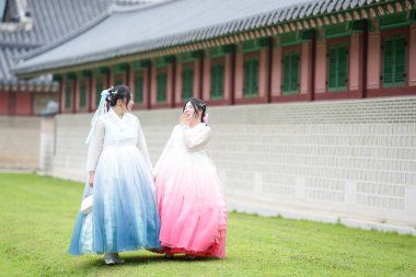 Yirmili yaşlarda iki Koreli kadın Gyeongbokgung Sarayı 'ndaki tarihi bir salonun önünde, açık bir yaz gökyüzü altında, pembe ve gökyüzü mavisi bir Hanbok giyip, çimlerin üzerinde yürüyorlar. Seul, Haziran.