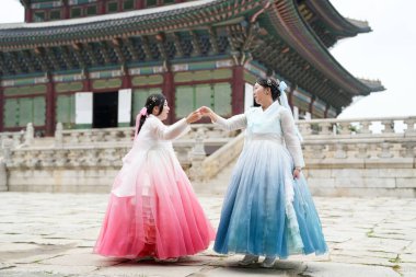 Yirmili yaşlardaki iki Koreli kadın, açık bir yaz gökyüzü altında güzel pembe ve gökyüzü mavisi Hanbok giyen tarihi mimarinin ortasında, Gyeongbokgung Sarayı 'nda güneşli bir plazada zarif bir şekilde dans ediyorlar. Seul, Haziran.