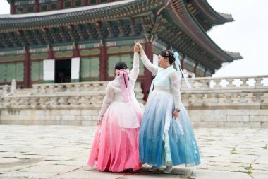 Yirmili yaşlardaki iki Koreli kadın, açık bir yaz gökyüzü altında güzel pembe ve gökyüzü mavisi Hanbok giyen tarihi mimarinin ortasında, Gyeongbokgung Sarayı 'nda güneşli bir plazada zarif bir şekilde dans ediyorlar. Seul, Haziran.