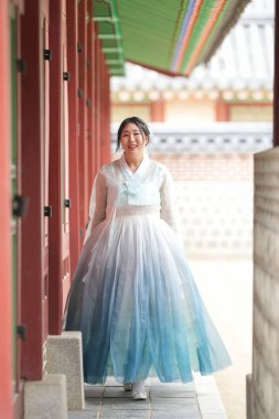 Yirmili yaşlarda Koreli bir kadın Gyeongbokgung Sarayı 'nın tarihi koridorunda yürüyor, açık bir gökyüzünün altında, süslü ahşap sütunlarla çerçevelenmiş, gökyüzü mavisi bir hanbok giyiyor. Seul, Haziran.
