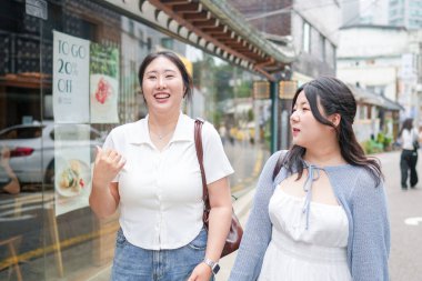 Süslü kafelerle dolu bir sokakta, yirmili yaşlarındaki iki Koreli kadın yumuşak bahar ışığı altında gündelik kıyafetlerle gezip sohbet ediyor, yollarını çerçeveleyen çiçekler açıyor. Seul, Jongno Bölgesi, bahar..