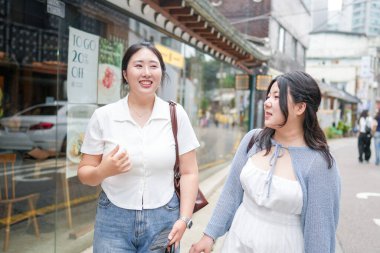 Süslü kafelerle dolu bir sokakta, yirmili yaşlarındaki iki Koreli kadın yumuşak bahar ışığı altında gündelik kıyafetlerle gezip sohbet ediyor, yollarını çerçeveleyen çiçekler açıyor. Seul, Jongno Bölgesi, bahar..
