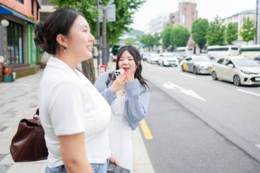 Hareketli bir bulvarda, yirmili yaşlardaki iki Koreli kadın otobüs durağında durup, yumuşak bahar ışığı altında otobüsü beklerken sessizce sohbet ediyorlar. Seul, Jongno Bölgesi, bahar..