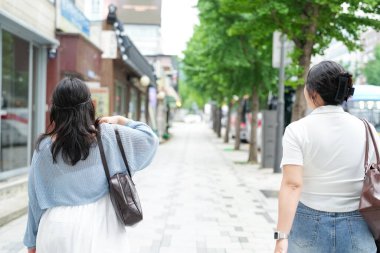 Süslü kafelerle dolu bir sokakta, yirmili yaşlarındaki iki Koreli kadın yumuşak bahar ışığı altında gündelik kıyafetlerle gezip sohbet ediyor, yollarını çerçeveleyen çiçekler açıyor. Seul, Jongno Bölgesi, bahar..