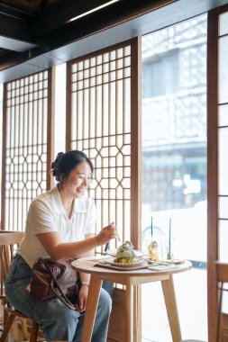 Pencere masasındaki şık bir kafede, yirmili yaşlardaki sıradan giyinmiş Koreli bir kadın yeşil bitkilerle çevrili yumuşak bahar ışığı altında lattenin yanında bir hamur işinin tadını çıkarıyor. Seul, Jongno Bölgesi, bahar..