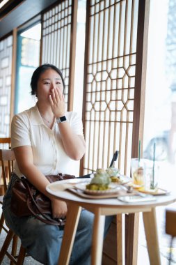 Pencere masasındaki şık bir kafede, yirmili yaşlardaki sıradan giyinmiş Koreli bir kadın yeşil bitkilerle çevrili yumuşak bahar ışığı altında lattenin yanında bir hamur işinin tadını çıkarıyor. Seul, Jongno Bölgesi, bahar..