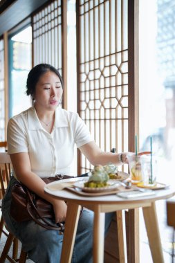 Pencere masasındaki şık bir kafede, yirmili yaşlardaki sıradan giyinmiş Koreli bir kadın yeşil bitkilerle çevrili yumuşak bahar ışığı altında lattenin yanında bir hamur işinin tadını çıkarıyor. Seul, Jongno Bölgesi, bahar..