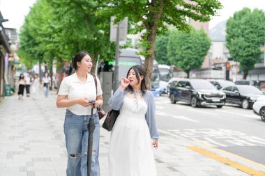 Süslü kafelerle dolu bir sokakta, yirmili yaşlarındaki iki Koreli kadın yumuşak bahar ışığı altında gündelik kıyafetlerle gezip sohbet ediyor, yollarını çerçeveleyen çiçekler açıyor. Seul, Jongno Bölgesi, bahar..