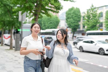 Süslü kafelerle dolu bir sokakta, yirmili yaşlarındaki iki Koreli kadın yumuşak bahar ışığı altında gündelik kıyafetlerle gezip sohbet ediyor, yollarını çerçeveleyen çiçekler açıyor. Seul, Jongno Bölgesi, bahar..
