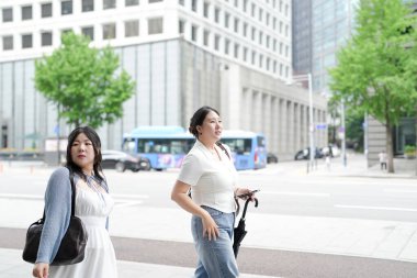 Yirmili yaşlardaki iki Koreli kadın sokak kaldırımında yürüyorlar, konuşuyorlar ve gülümsüyorlar şehrin aydınlık bahar ışığı altında uyanıp dükkanlardan geçerken. Seul, Jongno Bölgesi, bahar..
