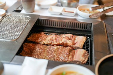 Bir kalp barbekü restoranında, bir sunucu yanan kömürlerin üzerinde bir et dilimi ızgara yapar, alevler, cızırdayan yağlardan yükselen buhar gibi kenarları yalar, havayı zengin aromalarla doldurur. Seul, Jung Bölgesi, bahar..