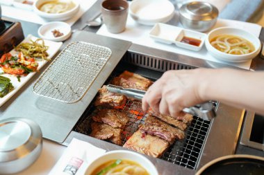 Bir kalp barbekü restoranında, bir sunucu yanan kömürlerin üzerinde bir et dilimi ızgara yapar, alevler, cızırdayan yağlardan yükselen buhar gibi kenarları yalar, havayı zengin aromalarla doldurur. Seul, Jung Bölgesi, bahar..