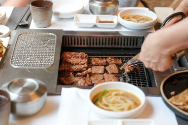 Bir kalp barbekü restoranında, bir sunucu yanan kömürlerin üzerinde bir et dilimi ızgara yapar, alevler, cızırdayan yağlardan yükselen buhar gibi kenarları yalar, havayı zengin aromalarla doldurur. Seul, Jung Bölgesi, bahar..