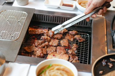 Bir kalp barbekü restoranında, bir sunucu yanan kömürlerin üzerinde bir et dilimi ızgara yapar, alevler, cızırdayan yağlardan yükselen buhar gibi kenarları yalar, havayı zengin aromalarla doldurur. Seul, Jung Bölgesi, bahar..