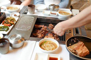 Bir kalp barbekü restoranında, bir sunucu yanan kömürlerin üzerinde bir et dilimi ızgara yapar, alevler, cızırdayan yağlardan yükselen buhar gibi kenarları yalar, havayı zengin aromalarla doldurur. Seul, Jung Bölgesi, bahar..