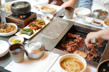 Bir kalp barbekü restoranında, bir sunucu yanan kömürlerin üzerinde bir et dilimi ızgara yapar, alevler, cızırdayan yağlardan yükselen buhar gibi kenarları yalar, havayı zengin aromalarla doldurur. Seul, Jung Bölgesi, bahar..