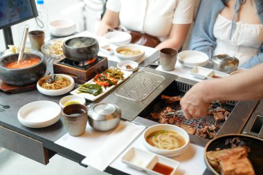 Bir kalp barbekü restoranında, bir sunucu yanan kömürlerin üzerinde bir et dilimi ızgara yapar, alevler, cızırdayan yağlardan yükselen buhar gibi kenarları yalar, havayı zengin aromalarla doldurur. Seul, Jung Bölgesi, bahar..