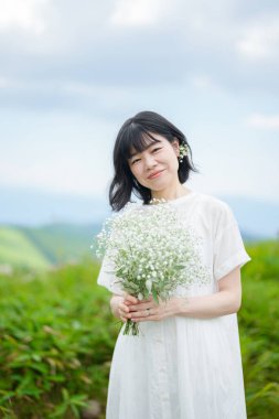 Beyaz elbiseli, orta boylu saçlı bir kadın açık gökyüzünün altındaki bir dağ yolunda tek başına dururken bebek nefesi tutuyor, bina yok. Nagano Bölgesi, Kirigamine, Japonya, yaz.