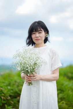Beyaz elbiseli, orta boylu saçlı bir kadın açık gökyüzünün altındaki bir dağ yolunda tek başına dururken bebek nefesi tutuyor, bina yok. Nagano Bölgesi, Kirigamine, Japonya, yaz.