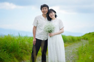 30 'lu yaşlarda bir çift dağ yolunda birlikte duruyor. Beyaz elbiseli orta boylu saçlı bir kadın, bebek nefesli bir kadın ve yanındaki adam. Nagano Bölgesi, Kirigamine, Japonya, yaz.