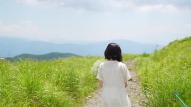 Arkadan, 30 'lu yaşlarda, beyaz elbiseli, orta boylu saçlı bir kadın bir dağ yolunda durup nefesini tutuyor.