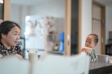 Çinli bir aile oturma odasında yemek yer, 30 'lu yaşlarda bir anne ve masada bir yaşındaki bebeği, mobilya ve ışığın arasında ekmek yiyen bir bebek. Bahar. Şangay 'da. Çin.