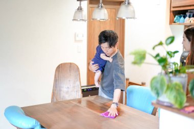 In a Mediterranean-style living room, a Japanese man in his 30s wipes the table with a cloth while holding a 3-month-old baby. Summer. Japan.