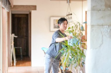 In a Mediterranean-style living room, a Japanese man in his 30s sprays water on a houseplant. Summer. Japan.