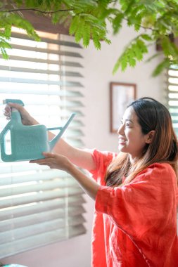 In a Mediterranean-style living room, a young Japanese woman in her 20s wearing an orange outfit sprays water on a houseplant. Japan. Summer.