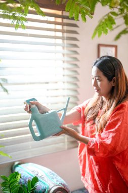 In a Mediterranean-style living room, a young Japanese woman in her 20s wearing an orange outfit sprays water on a houseplant. Japan. Summer.