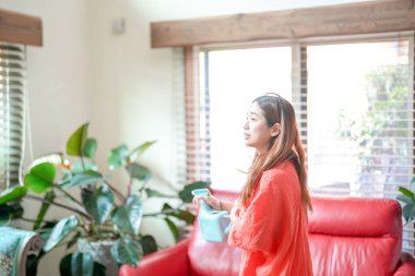 In a Mediterranean-style living room, a young Japanese woman in her 20s wearing an orange outfit sprays water on a houseplant. Japan. Summer.