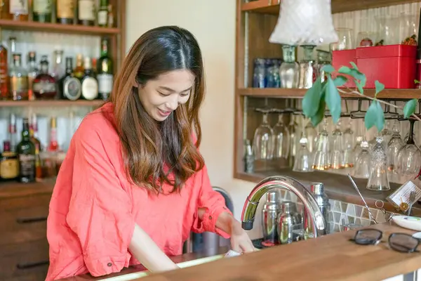 A young Japanese woman in her 20s wearing an orange outfit washing a wine glass in a stylish kitchen with many alcohol bottles. Japan. Summer.