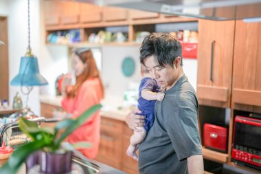 In a Mediterranean-style dining kitchen, a Japanese man in his 30s is holding a three-month-old baby while wiping the table with a cloth. Summer. Japan.