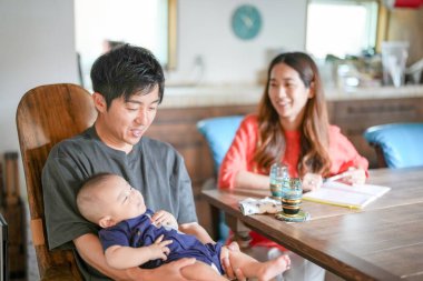 A man is holding a baby in his arms while a woman and a child sit at a table. The woman is smiling and the man is looking at the baby. The table is set with glasses and a book. The atmosphere is warm