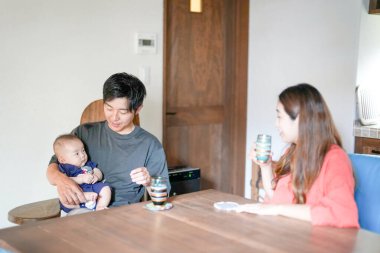 A Japanese father in his 30s holds a three-month-old baby while sitting at a Mediterranean-style dining table, sharing coffee and conversation with the mother in her 20s. Summer. Japan.