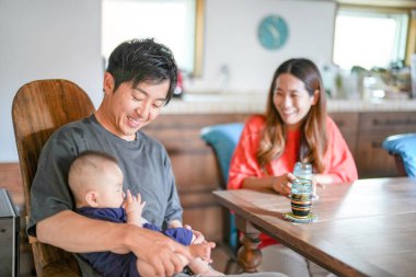A Japanese father in his 30s holds a three-month-old baby while sitting at a Mediterranean-style dining table, sharing coffee and conversation with the mother in her 20s. Summer. Japan.