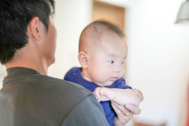 In a Mediterranean-style living room, a 3-month-old baby is in the arms of his Japanese father in his 30s. Summer. Japan.