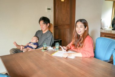 A man and a woman sit at a table with a baby in front of them. The woman is writing in a notebook while the man looks on. Scene is calm and peaceful, as the family enjoys a quiet moment together