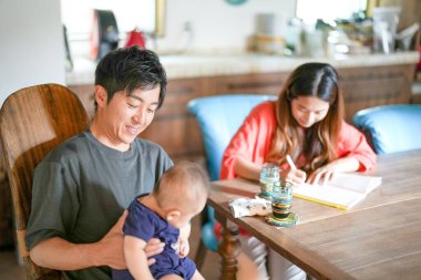 A man is holding a baby in a blue shirt. A woman is sitting at a table with a baby and a notebook
