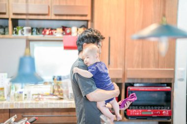 In a Mediterranean-style dining kitchen, a Japanese father in his 30s holds a three-month-old baby while wiping the table and talking to the mother in her 20s wearing orange clothes. Summer. Japan.