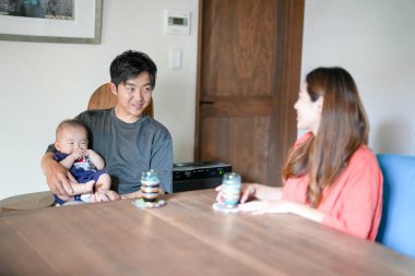 A Japanese father in his 30s holds a three-month-old baby while sitting at a Mediterranean-style dining table, sharing coffee and conversation with the mother in her 20s. Summer. Japan.