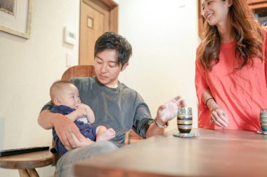 A Japanese father in his 30s holds a three-month-old baby while sitting at a Mediterranean-style dining table, sharing coffee and conversation with the mother in her 20s. Summer. Japan.