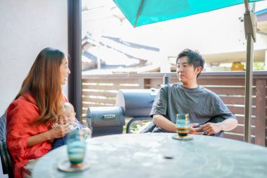 A man and a woman are sitting at a table with a baby. The man is holding a cup and the woman is holding a baby. Scene is warm and friendly