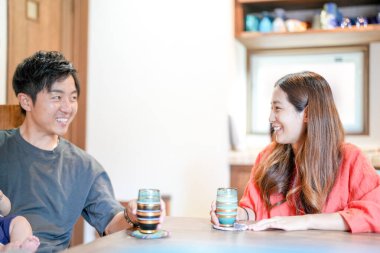 A Japanese father in his 30s holds a three-month-old baby while sitting at a Mediterranean-style dining table, sharing coffee and conversation with the mother in her 20s. Summer. Japan.