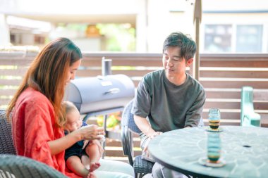 A man and a woman are sitting at a table with a baby in front of them. The man is holding the baby while the woman is feeding the baby. Scene is warm and loving